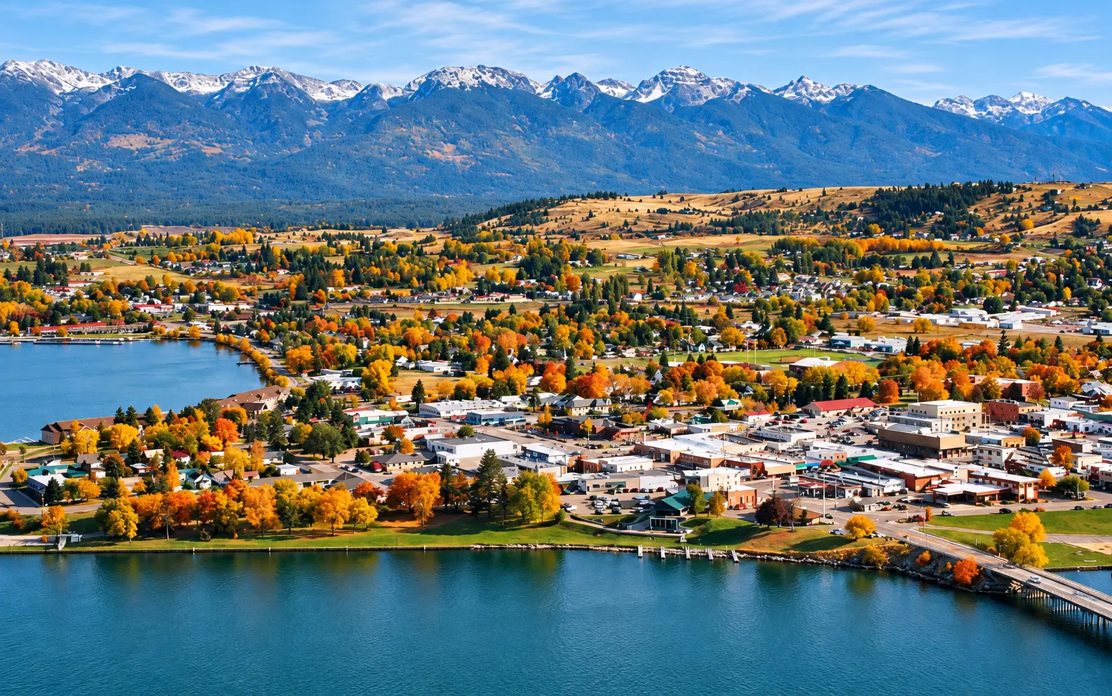 Elevated view over Polson, Montana, with autumn-colored trees across the lakefront town, the Mission Mountains rising behind, and a bridge crossing the water in the foreground