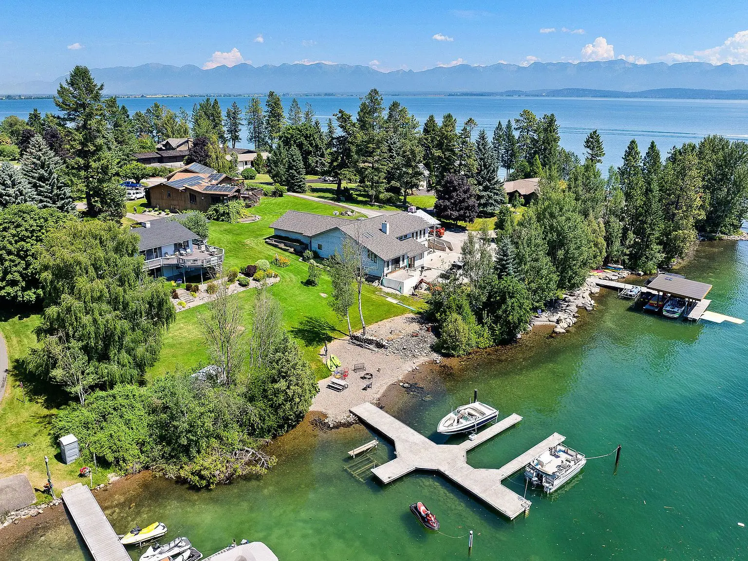 Aerial view of lakefront homes with private docks on clear turquoise water, backed by green lawns and pines, with hazy mountains across Flathead Lake near Lakeside, Montana
