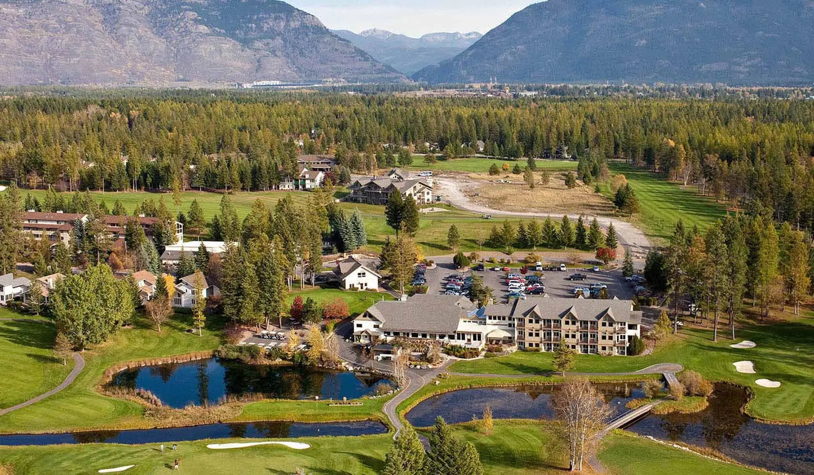 A golf course and stone-and-timber lodge seen from above, ringed by pine forest with hazy mountains behind, near Columbia Falls, Montana